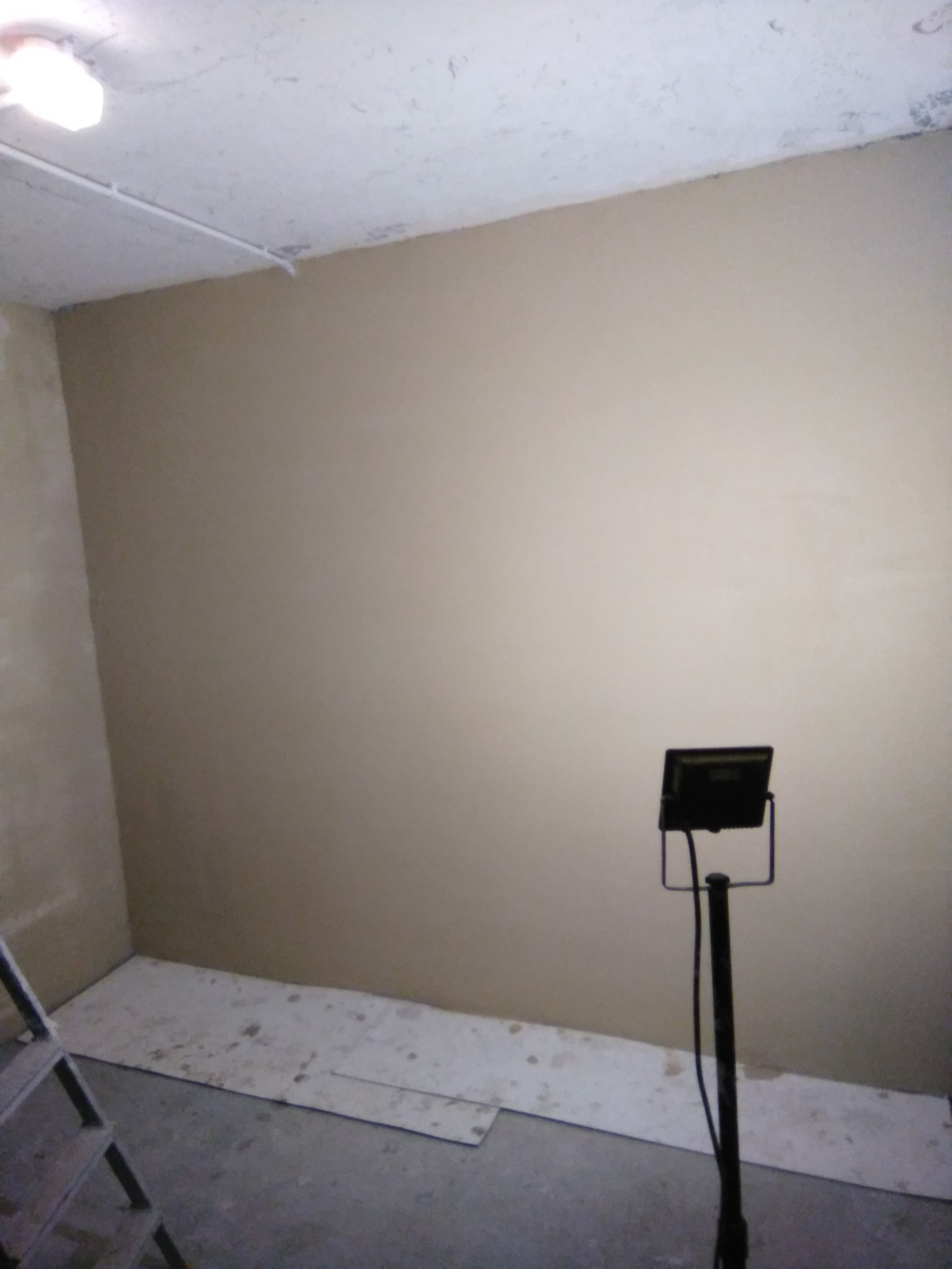 Świeżo pomalowana ściana w pokoju, zabezpieczona podłoga, widoczna drabina i lampa halogenowa na statywie.