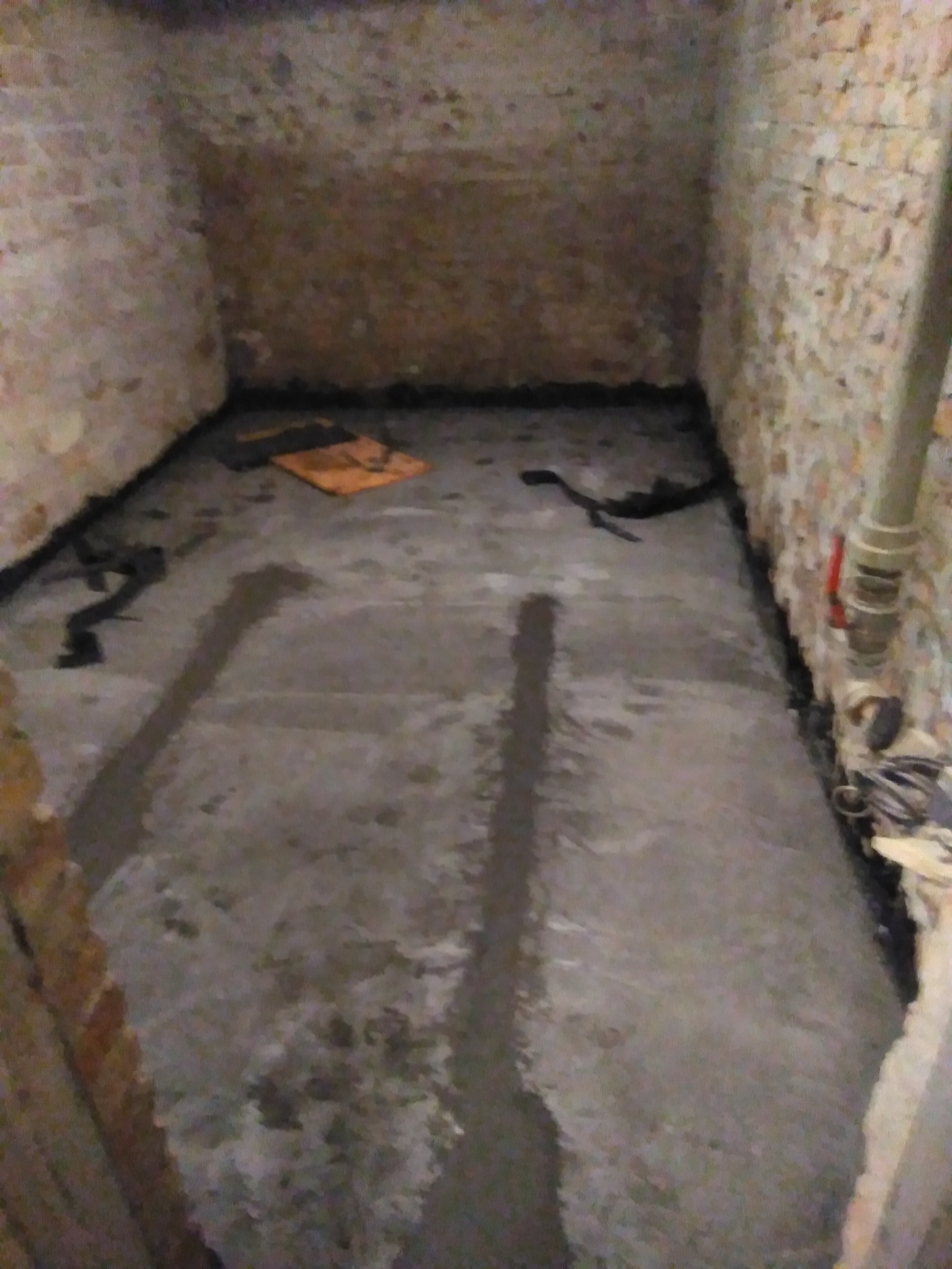 Piwnica w trakcie hydroizolacji: świeżo wylany beton na podłodze, uszczelnienie przy ścianach z cegły, widoczny fragment rury i narzędzia.