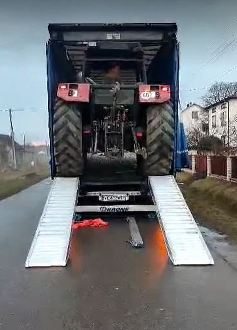 Czerwony traktor wjeżdża po rampie na platformę ciężarówki z otwartą plandeką, widok z tyłu, mokra nawierzchnia.