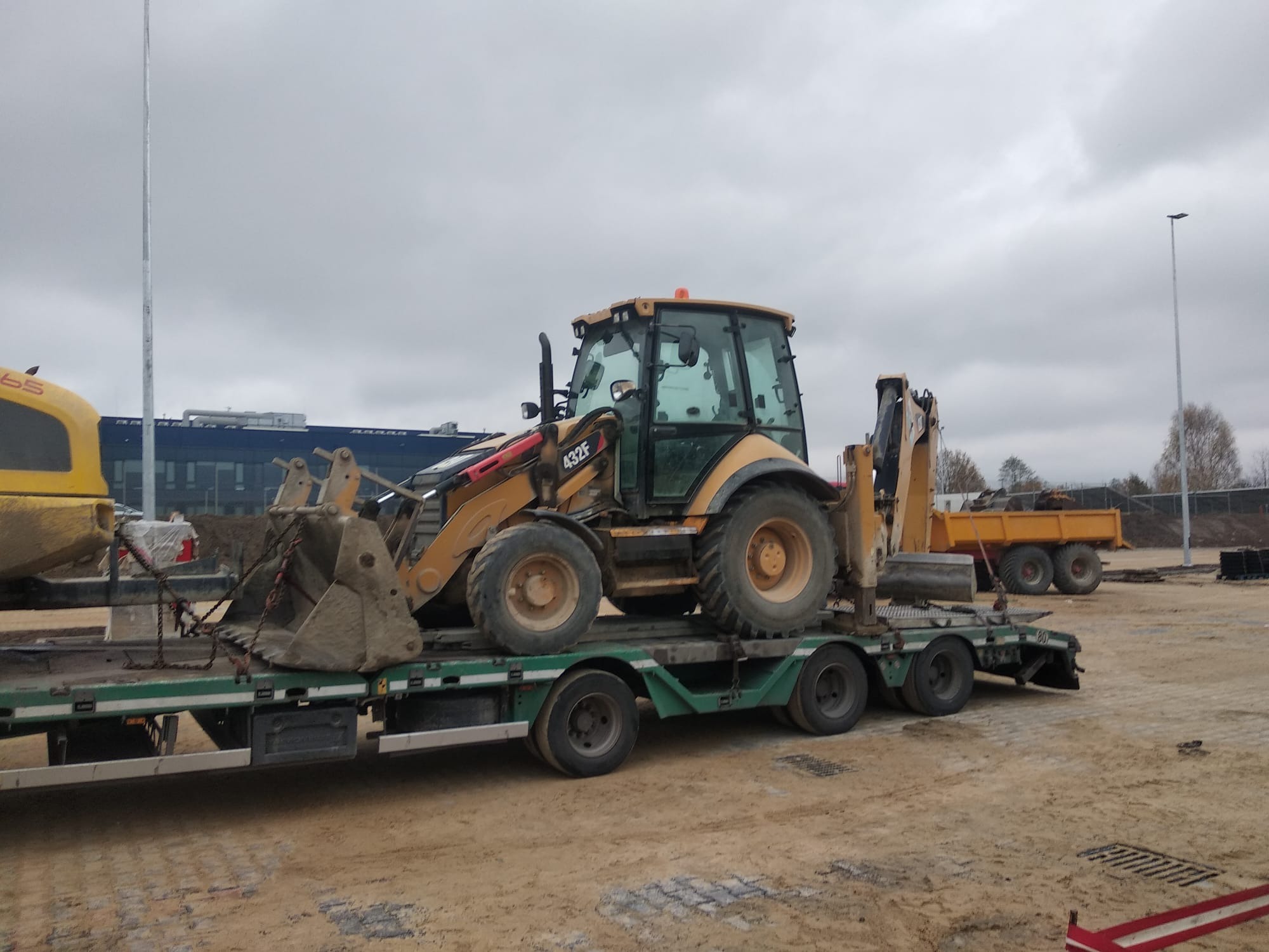 Żółta koparko-ładowarka Case 432F zabezpieczona łańcuchami na zielonej niskopodwoziowej platformie transportowej, z widoczną przyczepą wywrotką w tle, na placu budowy w pochmurny dzień.
