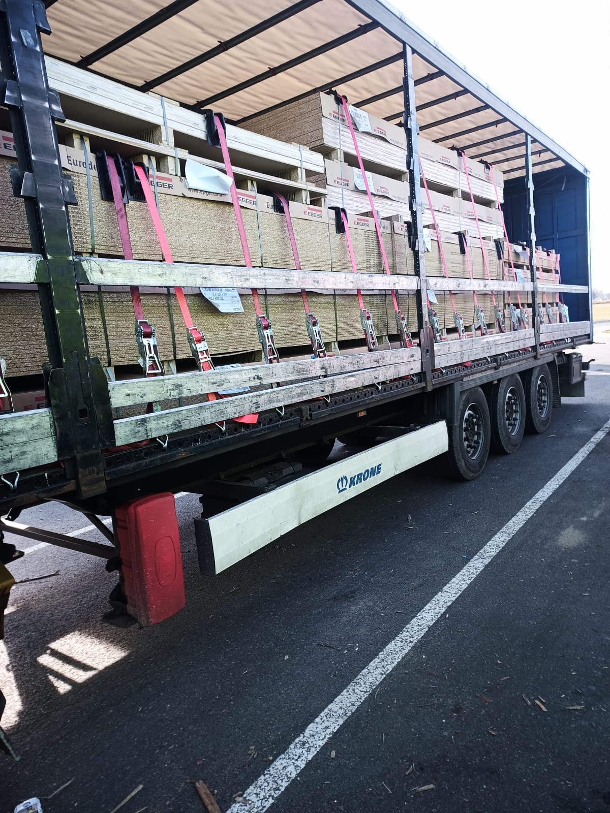 Ciężarówka z otwartą naczepą załadowana paletami z płytami drewnopochodnymi, zabezpieczonymi czerwonymi pasami, widoczna marka Krone na osłonie podwozia.