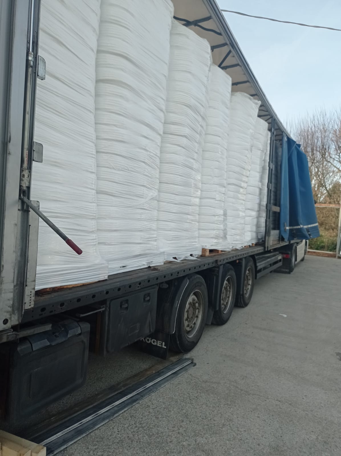 Ciężarówka z naczepą marki Kogel załadowana paletami z workami, zabezpieczonymi białą folią, widok z boku na koła i podwozie, niebieska plandeka z tyłu.