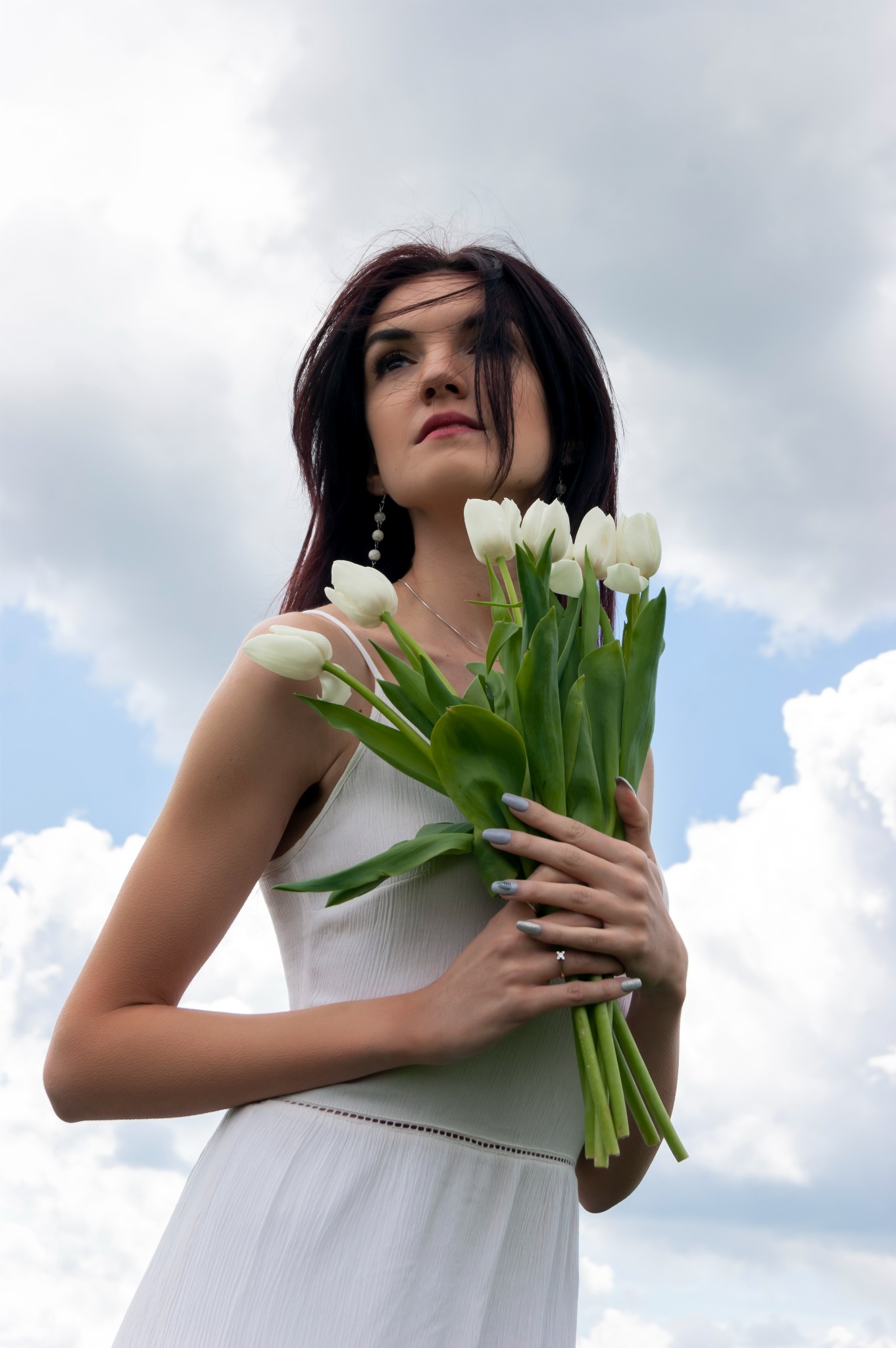 Kobieta w białej sukience trzyma bukiet białych tulipanów na tle błękitnego nieba z chmurami, spoglądając w górę.