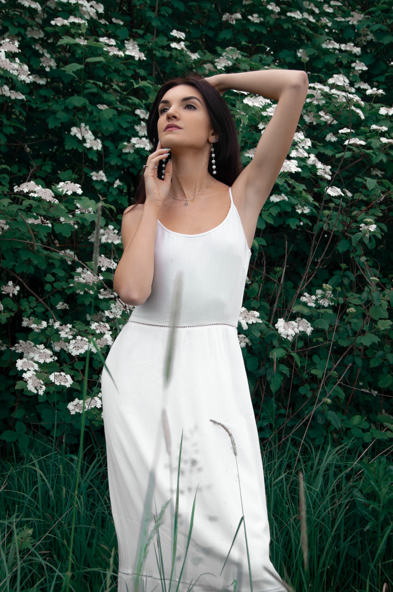 Kobieta w białej sukience, z długimi kolczykami i naszyjnikiem, pozuje na tle kwitnącego krzewu.