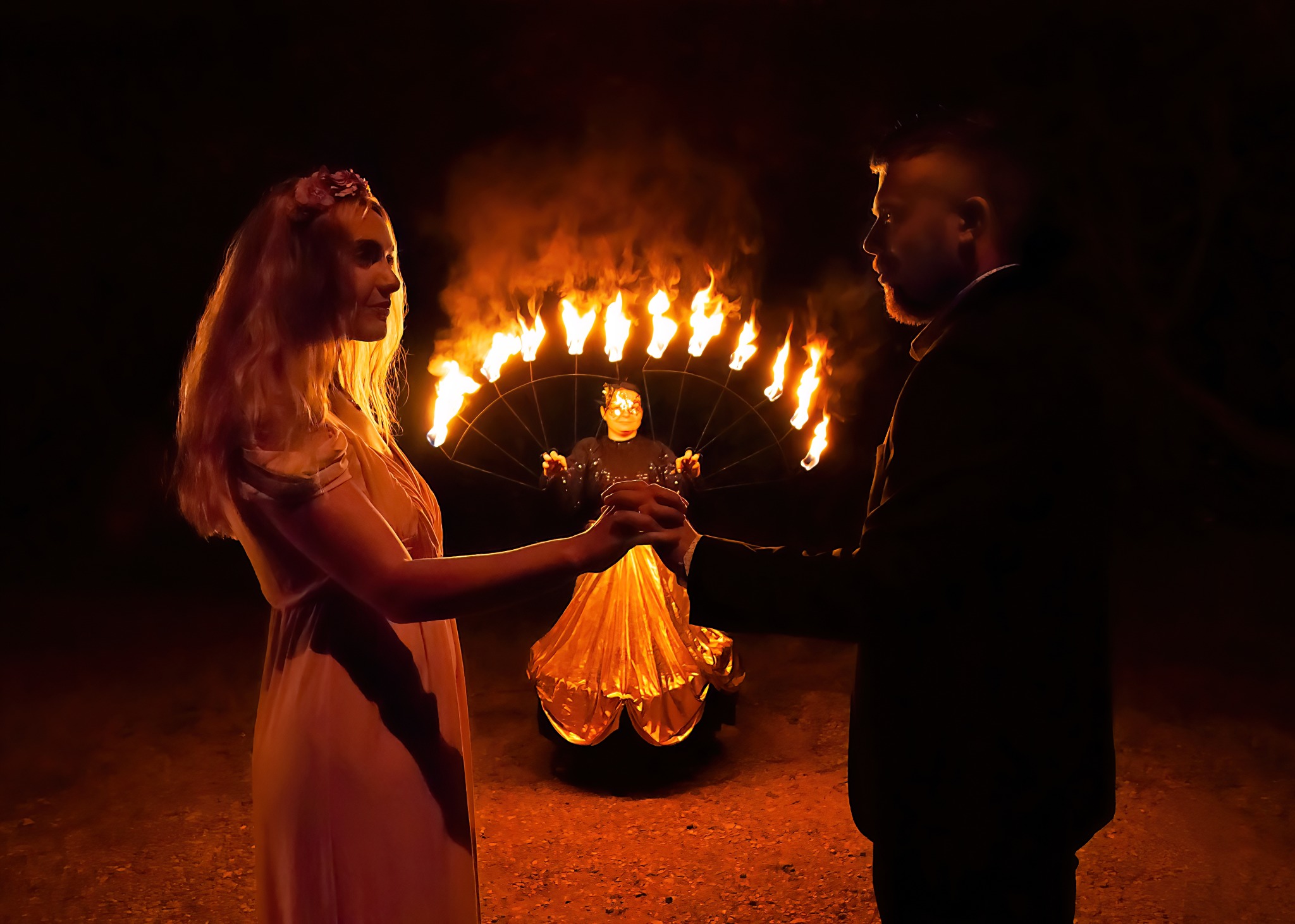 Para trzymająca się za ręce w ciemności, oświetlona przez pokaz ognia za nimi; kobieta w jasnej sukni z kwiatami we włosach, mężczyzna w ciemnym garniturze, w tle osoba z wachlarzem płonących...