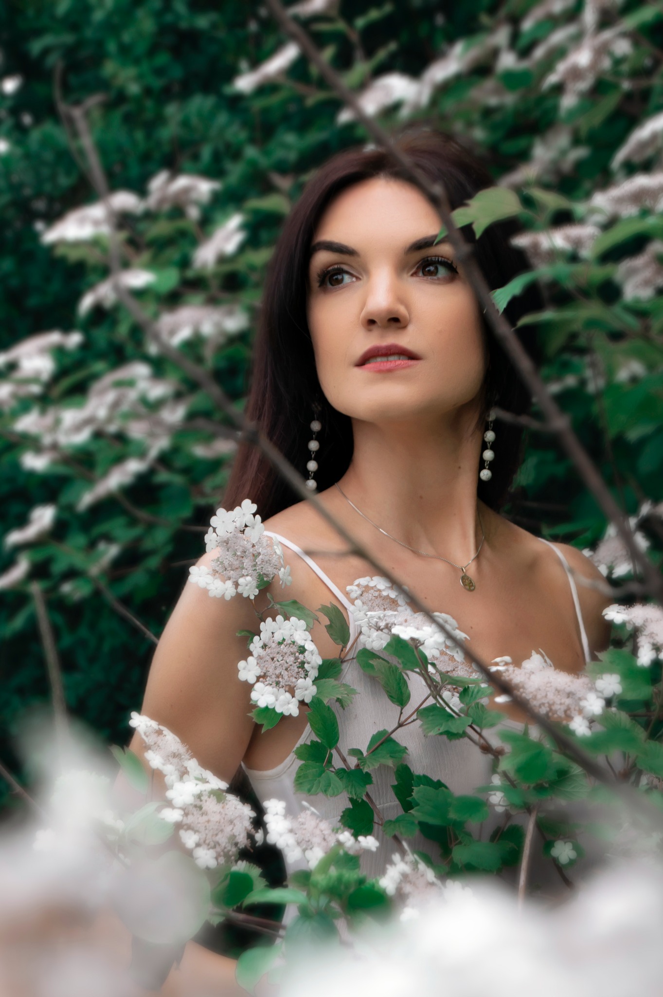 Portret kobiety w zwiewnej białej sukience, otoczonej kwitnącymi gałązkami i zielenią, z długimi kolczykami i delikatnym naszyjnikiem, wykonany w Bielsku-Białej, spoglądającej w dal.