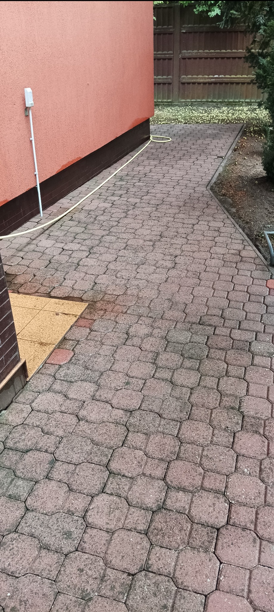 Czerwona kostka brukowa na podjeździe przed domem, widoczny fragment elewacji w kolorze łososiowym i biały kabel biegnący wzdłuż ściany, oraz częściowo umyta, jaśniejsza powierzchnia kostki.