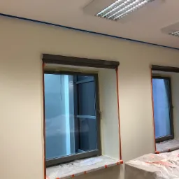 Przygotowanie do malowania biura: świeżo pomalowane ściany w odcieniach beżu, okna zabezpieczone taśmą malarską, widoczne zabezpieczone meble folią, niebieska linia taśmy na suficie.