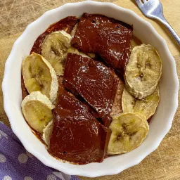 Deser w białej miseczce: czekoladowe bloki z wytłoczonym wzorem i suszone plasterki banana, na drewnianym tle z fioletową ściereczką w białe kropki i widelcem.