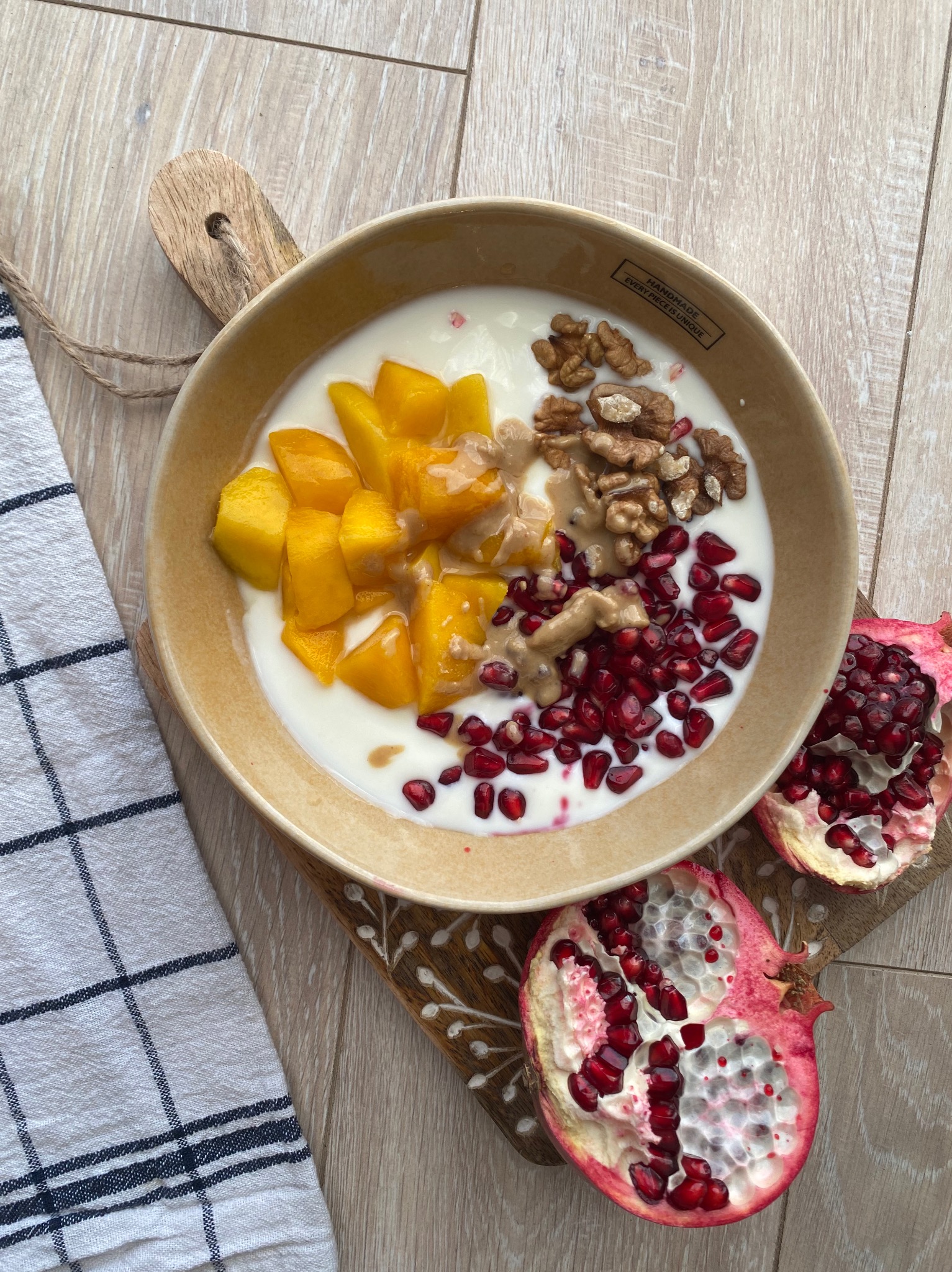 Miska z jogurtem, mango, orzechami włoskimi, sosem tahini i granatem na drewnianej desce obok lnianej ściereczki w kratę.