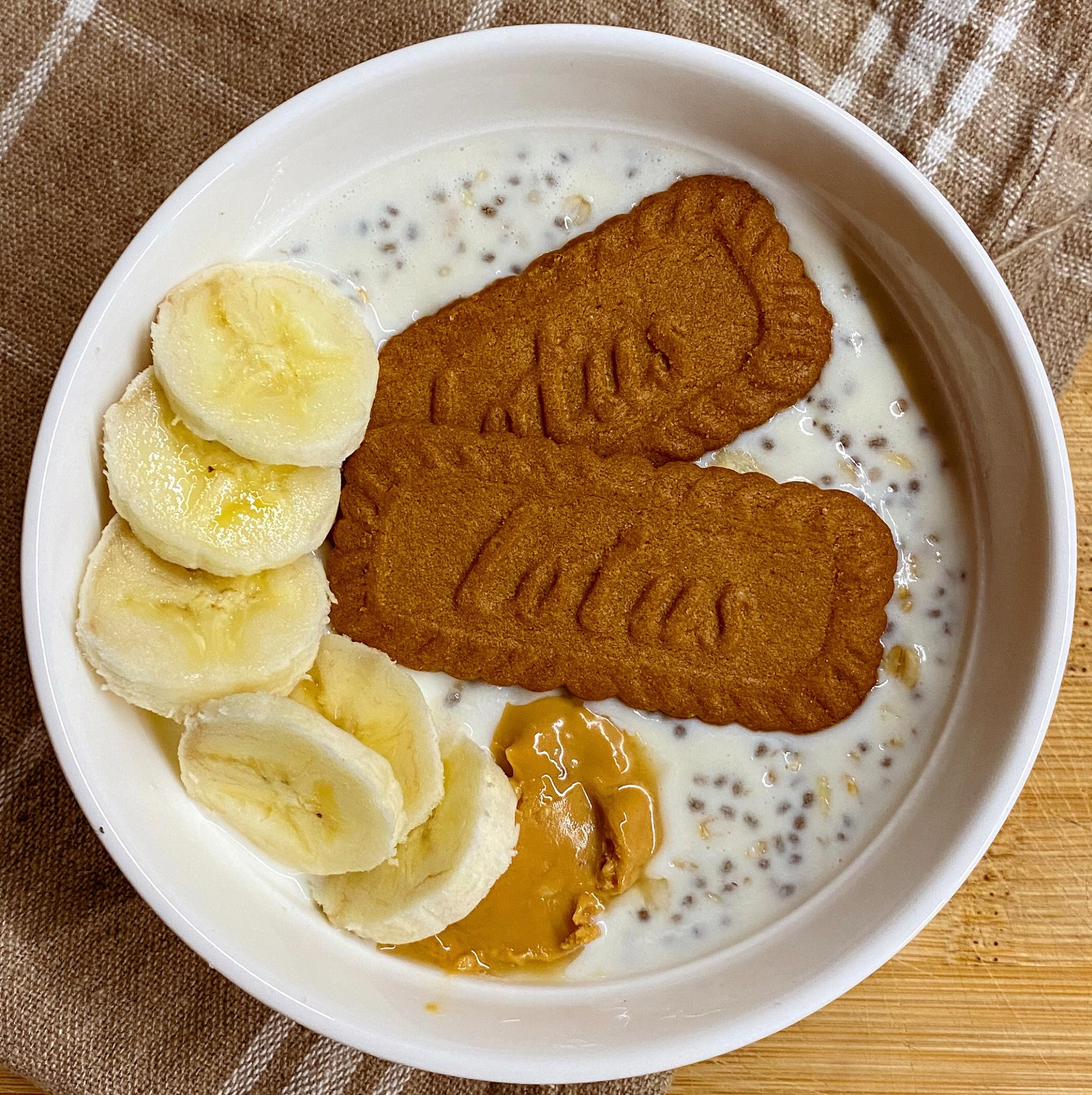 Śniadaniowa miska z mlekiem roślinnym, nasionami chia, bananem, masłem orzechowym i dwoma ciasteczkami Lotus Biscoff na drewnianym blacie.