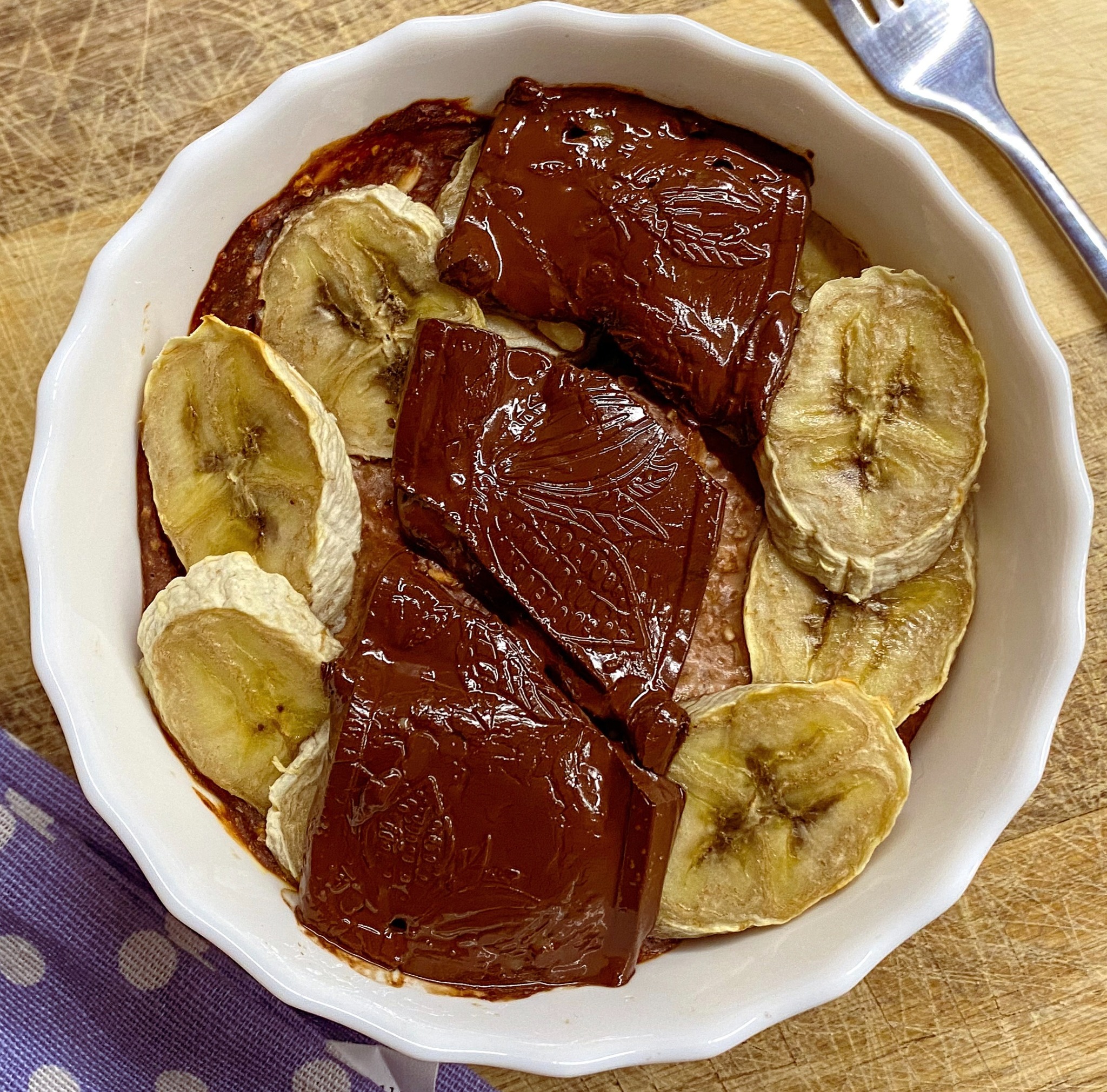 Deser w białej miseczce: czekoladowe bloki z wytłoczonym wzorem i suszone plasterki banana, na drewnianym tle z fioletową ściereczką w białe kropki i widelcem.