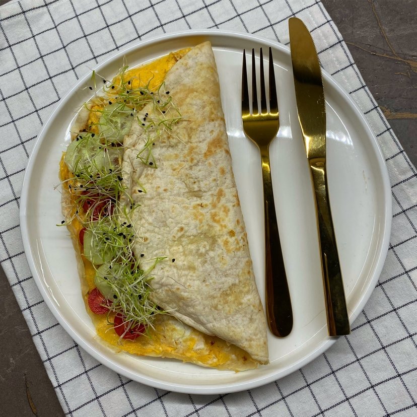 Tortilla wrap z jajkiem, warzywami i kiełkami na białym talerzu z złotymi sztućcami, na tle szarego blatu i białej ściereczki w czarną kratę.