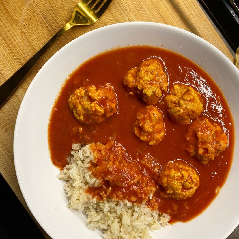 Porcja pulpetów w sosie pomidorowym z ryżem brązowym, podana na białym talerzu, z widocznym złotym widelcem na drewnianym tle.