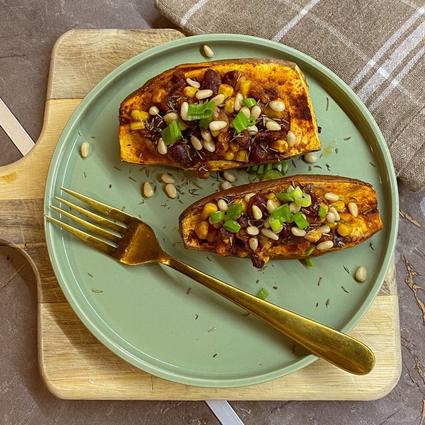 Dwa pieczone bataty przekrojone na pół, wypełnione farszem z kukurydzy, fasoli i orzeszków pinii, posypane zielonym szczypiorkiem, podane na zielonym talerzu z pozłacanym widelcem, na drewnianej...