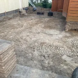 Przygotowany teren pod budowę z widocznym fundamentem budynku, stosem płyt chodnikowych, oponami i małą koparką w tle.