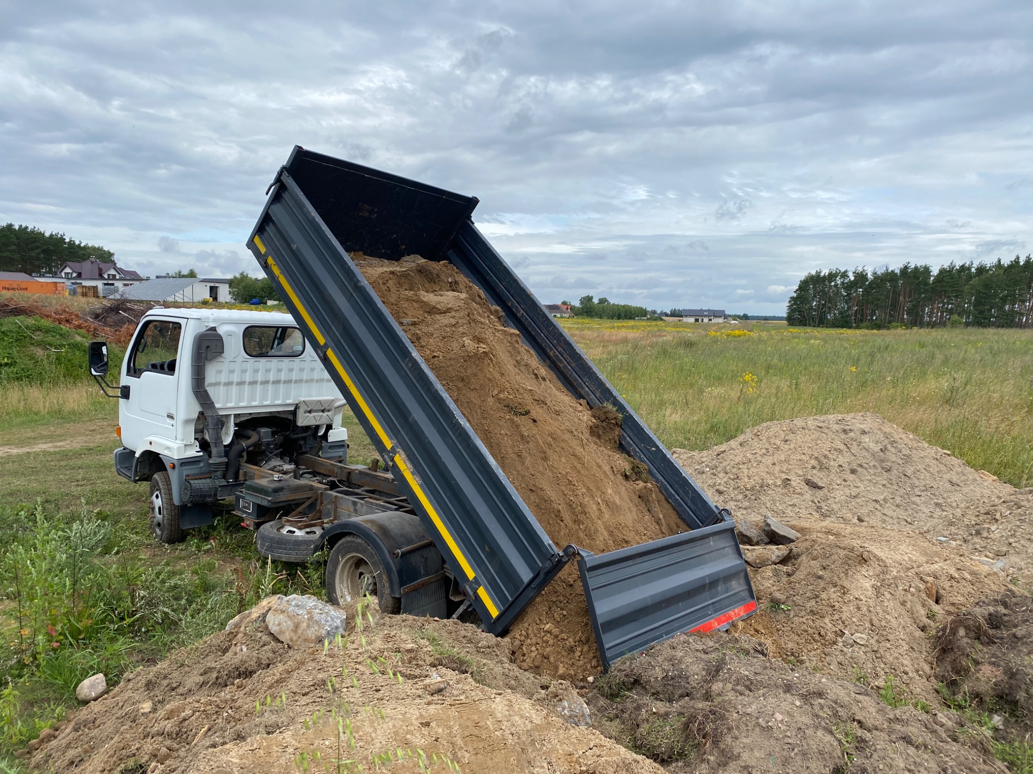 Mała, biała wywrotka z podniesioną skrzynią ładunkową, wysypująca ziemię na pryzmę. W tle widoczne zabudowania mieszkalne i łąka pod pochmurnym niebem.