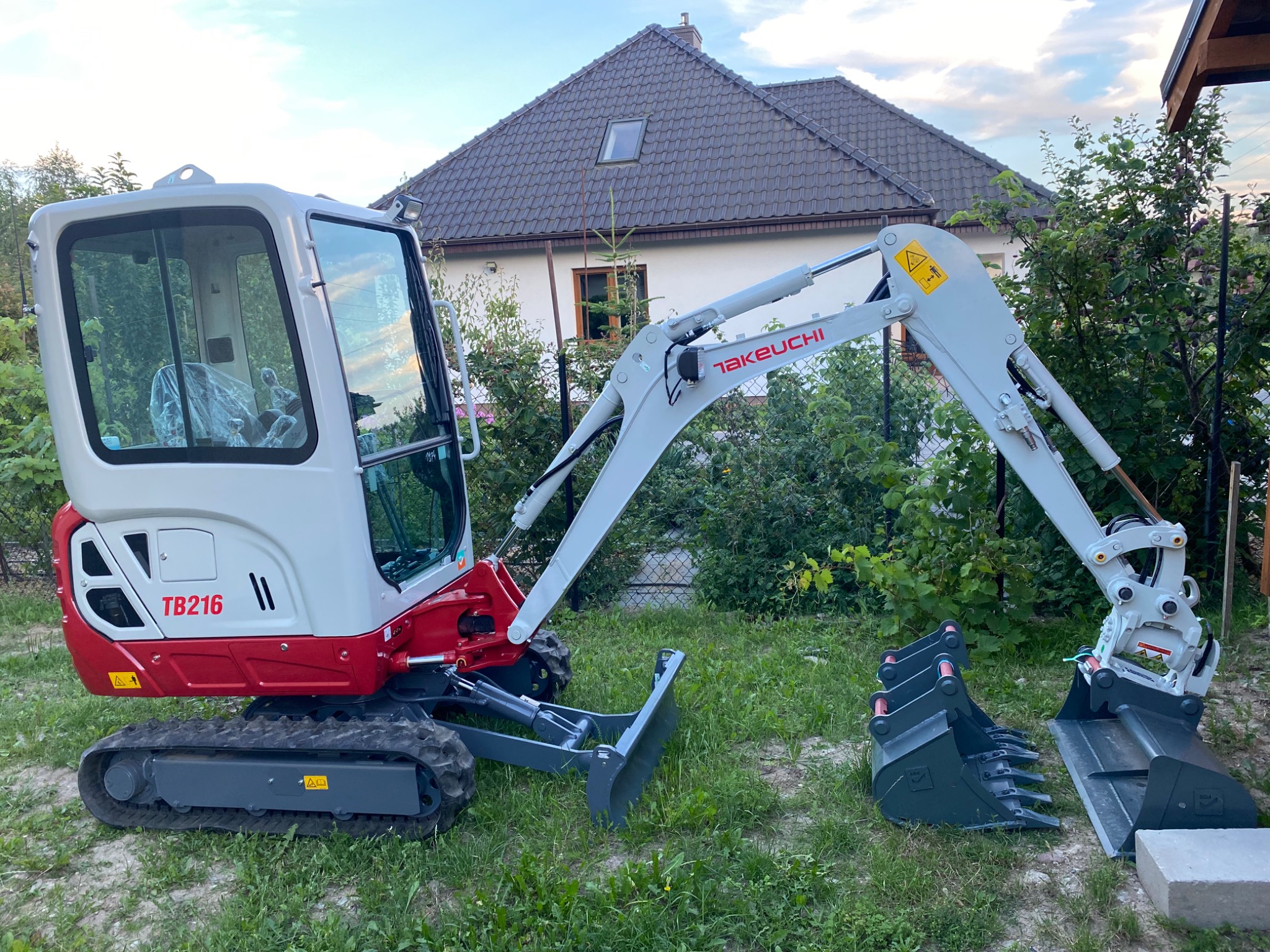Nowa, biało-czerwona minikoparka Takeuchi TB216 z trzema łyżkami o różnych szerokościach leżącymi obok, na trawiastym terenie przed domem jednorodzinnym.