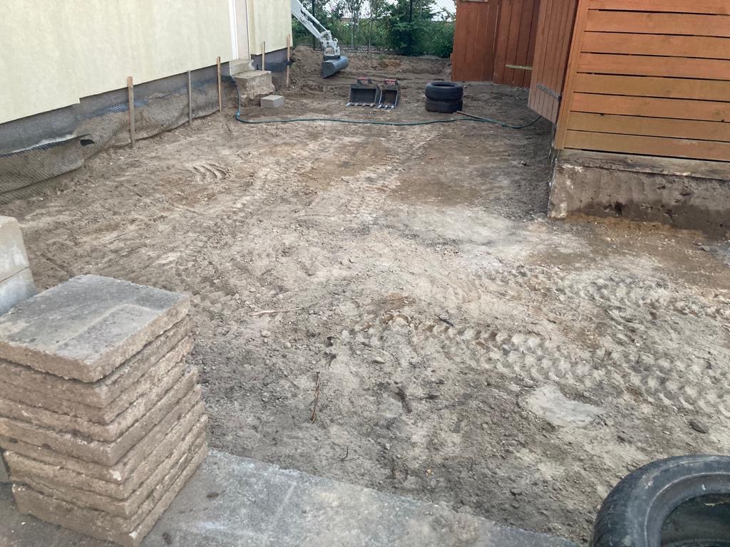 Przygotowany teren pod budowę z widocznym fundamentem budynku, stosem płyt chodnikowych, oponami i małą koparką w tle.