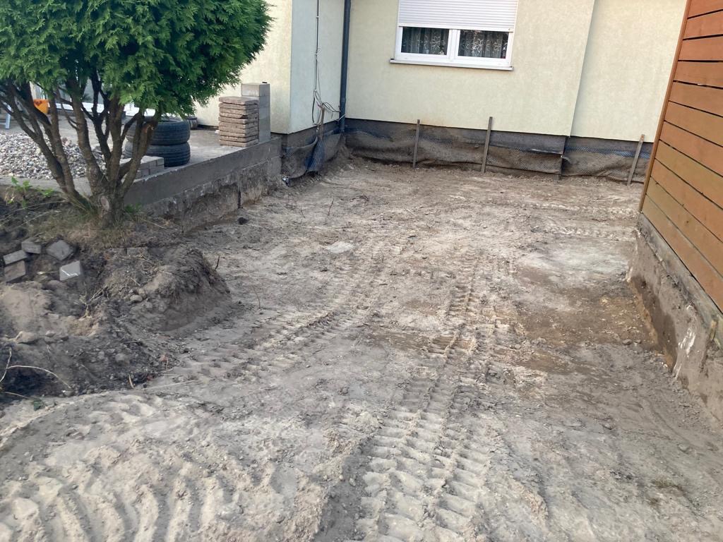 Przygotowany teren pod budowę w ciasnej zabudowie, widoczne ślady opon po maszynach, częściowo odsłonięte fundamenty budynku i ogrodzenie z desek.