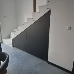 Nowoczesne schody wspornikowe z zabudową pod schodami w postaci ciemnych szafek, widoczne z boku w jasnym wnętrzu domu.