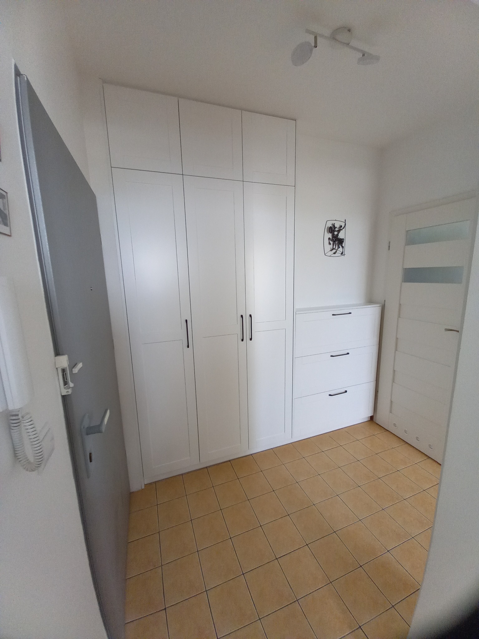 Biała szafa na wymiar z trzema segmentami drzwi i komodą z szufladami, uzupełniająca aranżację przedpokoju z brązową podłogą i szarą ścianą z domofonem.