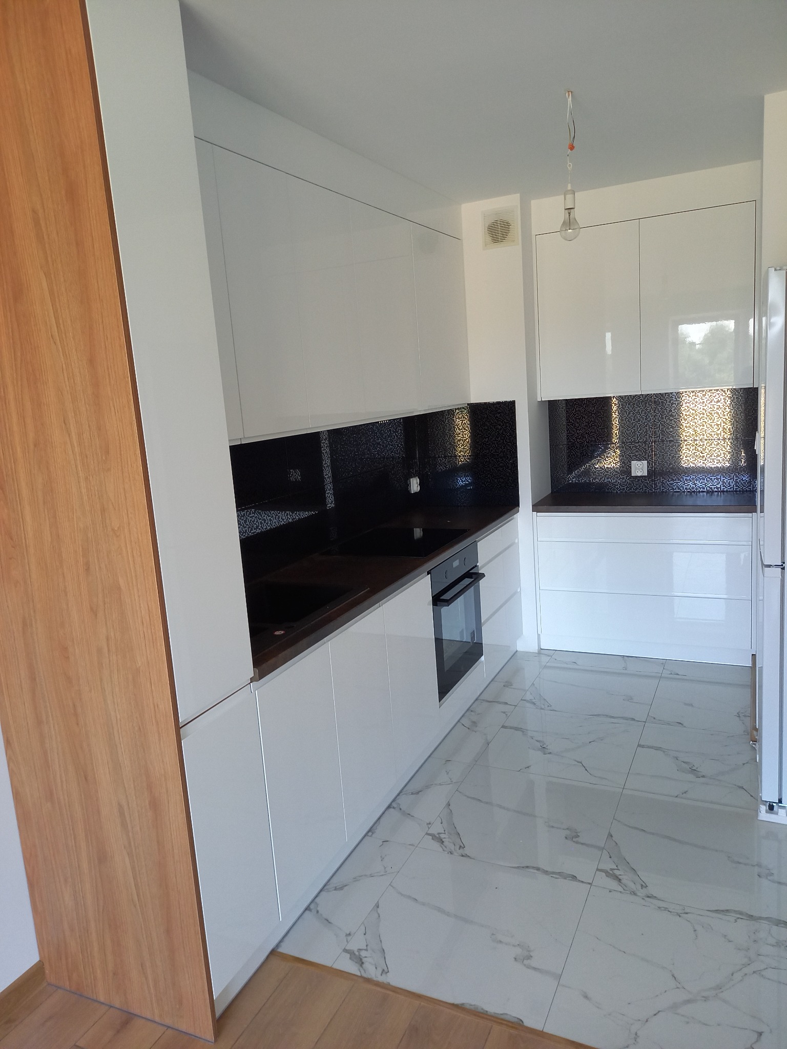 Nowoczesna, biała kuchnia z czarnym panelem nad blatem i marmurową podłogą, widok na zabudowę z piekarnikiem i niezabezpieczoną żarówką wiszącą z sufitu.