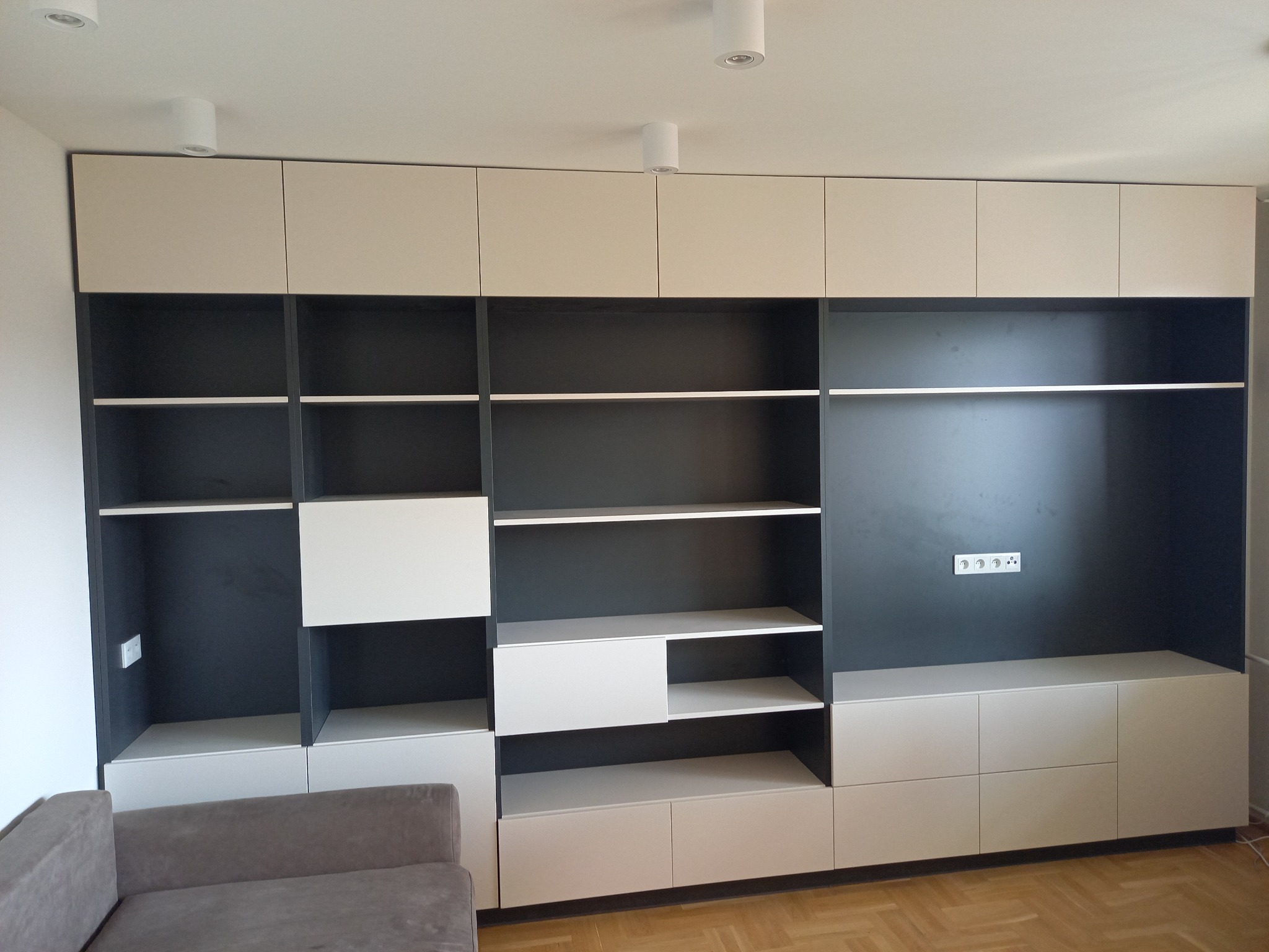 Nowoczesna zabudowa meblowa w salonie, z elementami otwartych półek w kolorze grafitowym i zamkniętych szafek w odcieniach beżu, uzupełniona o minimalistyczne oświetlenie sufitowe i fragment sofy...