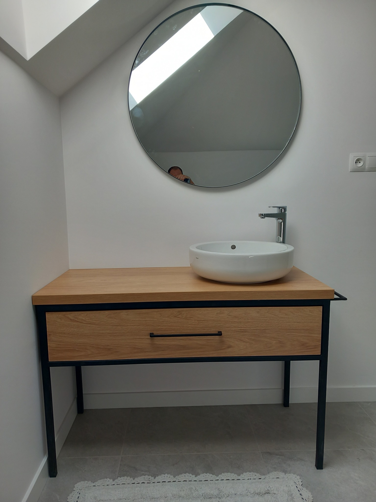 Biała umywalka nablatowa na drewnianym blacie z szufladą i czarną metalową ramą, okrągłe lustro na ścianie w łazience na poddaszu.