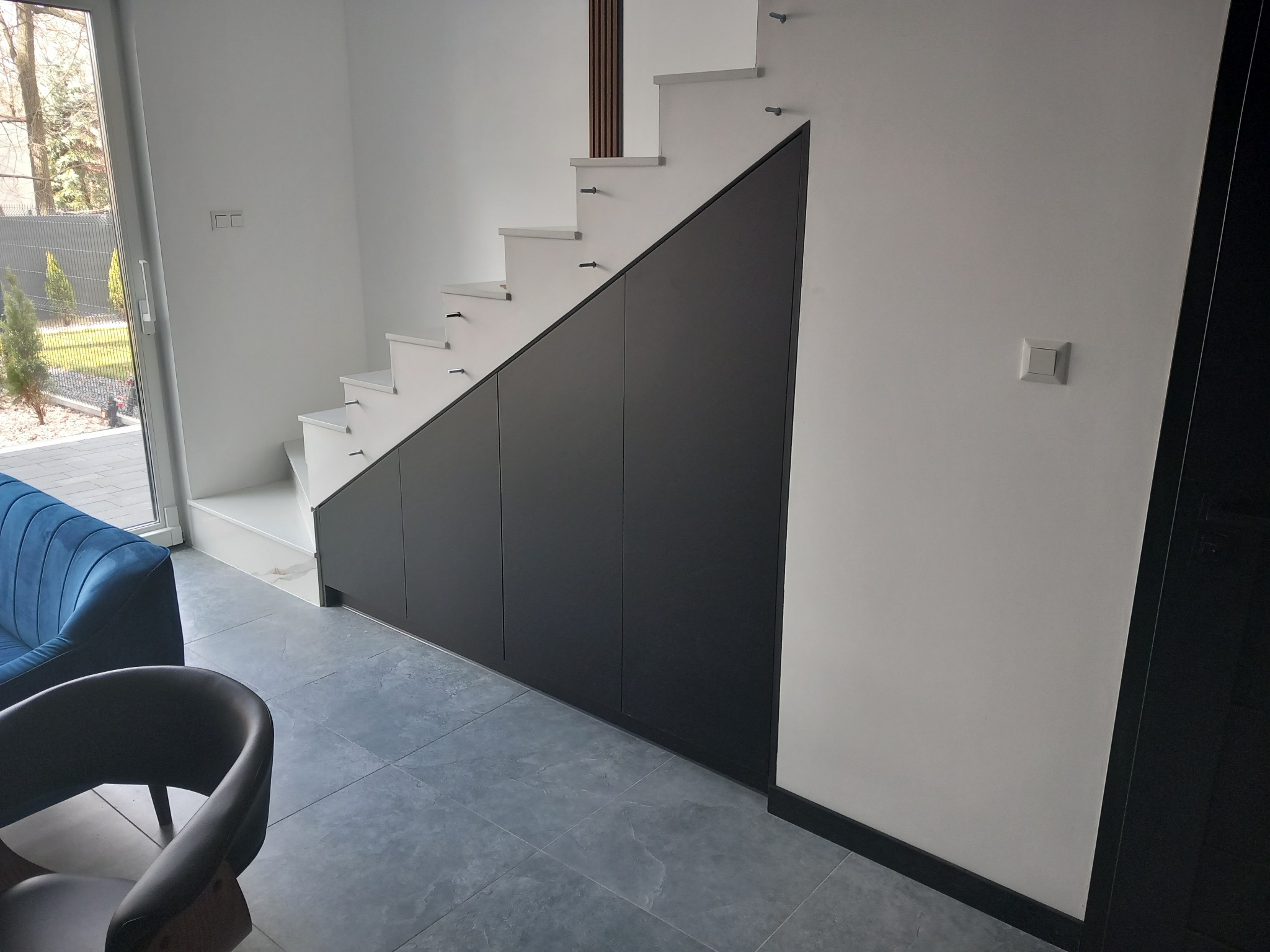 Nowoczesne schody wspornikowe z zabudową pod schodami w postaci ciemnych szafek, widoczne z boku w jasnym wnętrzu domu.