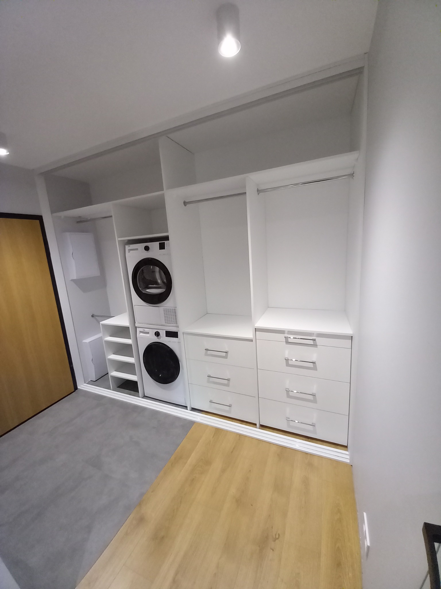 Biała szafa wnękowa z pralką i suszarką w zabudowie, widoczne szuflady z chromowanymi uchwytami, półki oraz drążek na ubrania, na podłodze panele w dwóch odcieniach szarości i beżu.