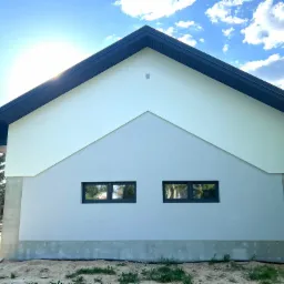 Fragment elewacji domu jednorodzinnego z dwoma oknami, widoczne połączenie dwóch odcieni szarości i białego, z czarnym dachem i elementami wykończeniowymi, na tle błękitnego nieba z chmurami.