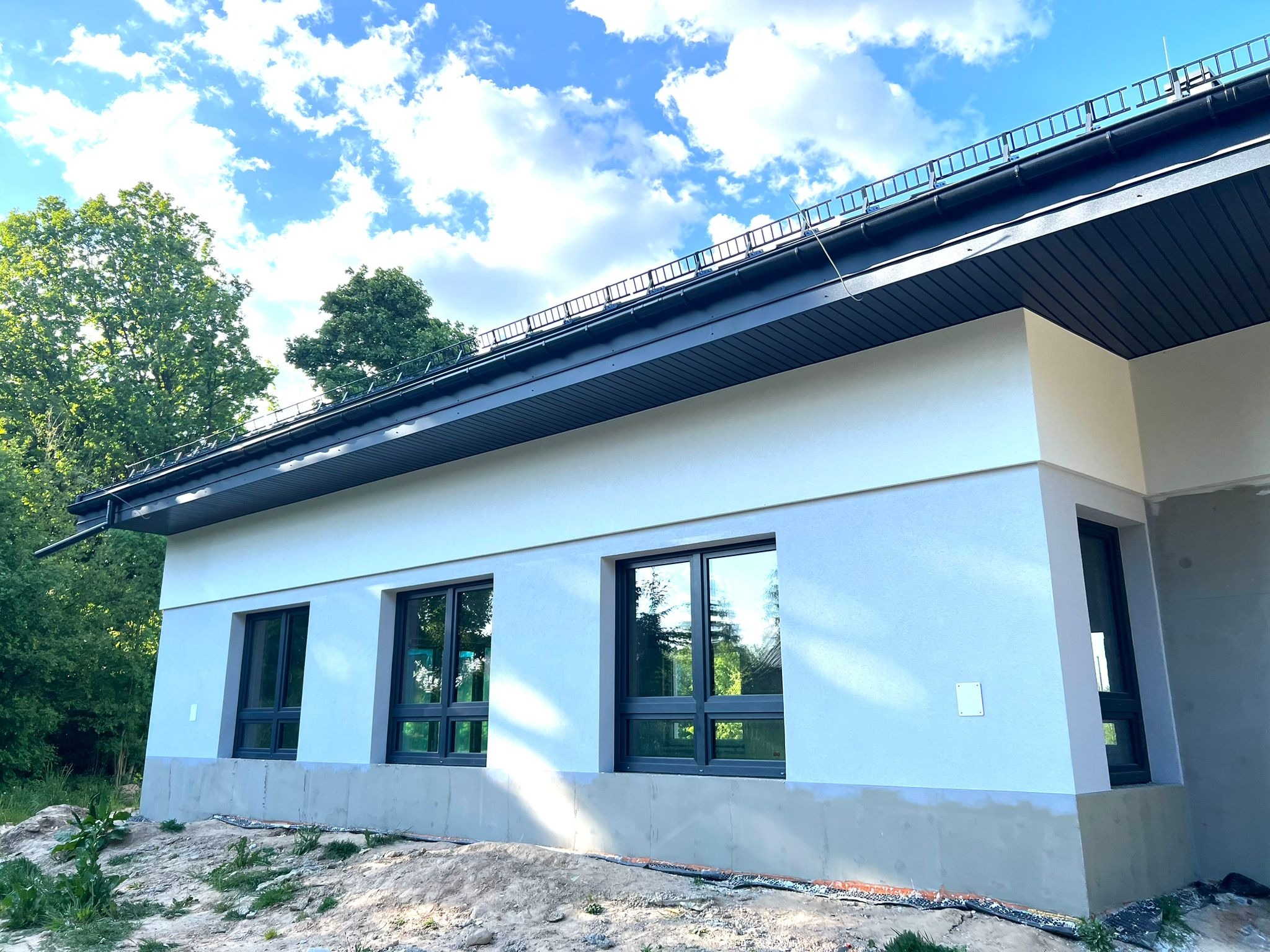 Nowoczesny dom jednorodzinny w trakcie wykańczania elewacji, widoczne trzy okna z ciemnymi ramami, jasna elewacja i szara podmurówka, na dachu zainstalowane zabezpieczenia przeciwśniegowe, w tle...