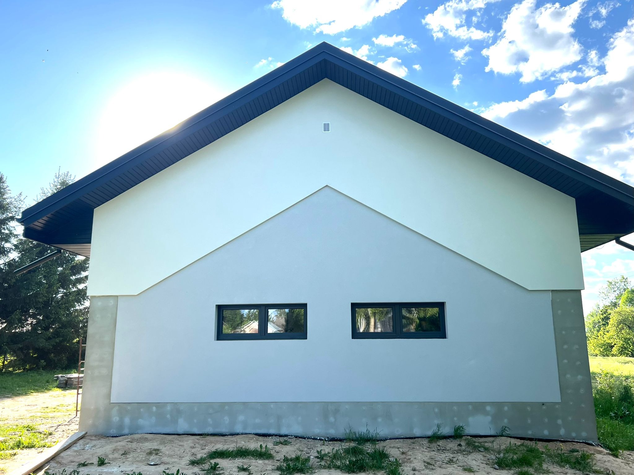 Fragment elewacji domu jednorodzinnego z dwoma oknami, widoczne połączenie dwóch odcieni szarości i białego, z czarnym dachem i elementami wykończeniowymi, na tle błękitnego nieba z chmurami.