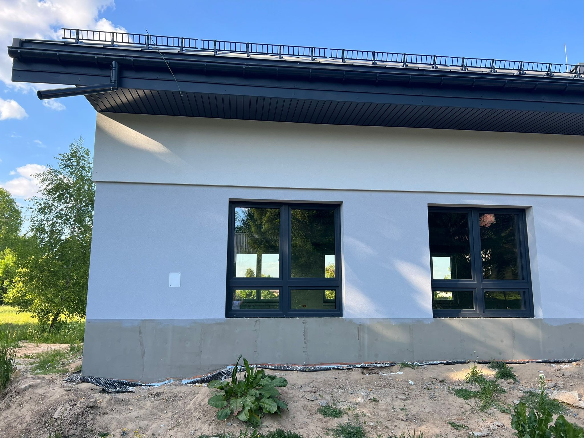 Fragment ściany budynku z dwoma oknami, elewacja w jasnym odcieniu szarości, cokół w szarym betonie, widoczne prace wykończeniowe wokół budynku.