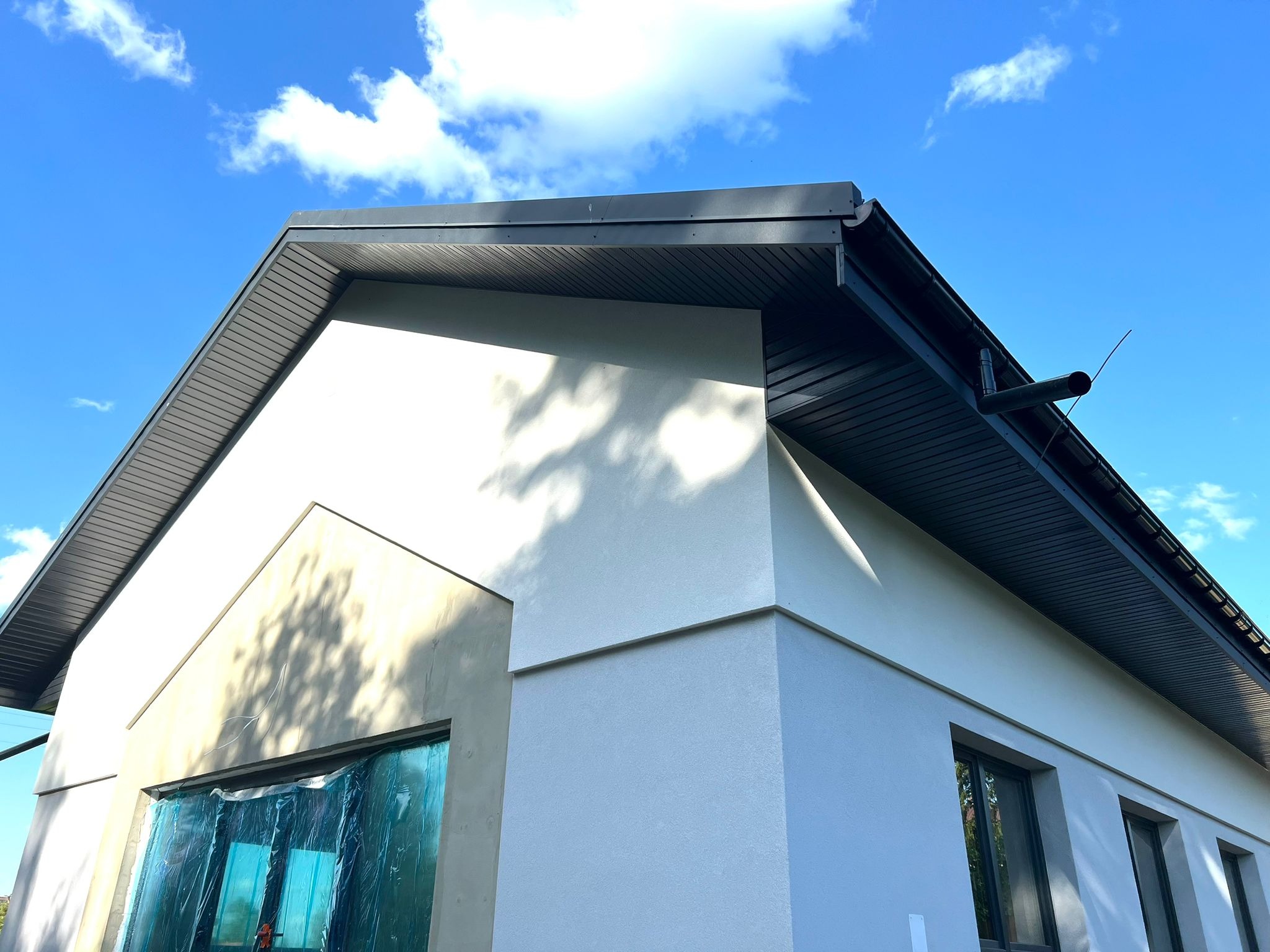 Fragment elewacji budynku z nową, jasną strukturą i ciemnym dachem, okna zabezpieczone folią, widoczne fragmenty instalacji odprowadzającej wodę deszczową, na ścianie cień drzewa.