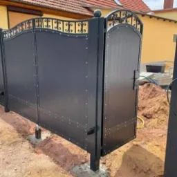 Czarna brama wjazdowa z elementami kutymi, widoczne prace ziemne w trakcie montażu, automat do bramy w tle.