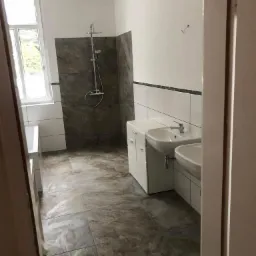 Nowoczesna łazienka z dwoma umywalkami na białych szafkach, szarymi płytkami pod prysznicem i na podłodze, oraz białymi płytkami z czarnym paskiem na ścianach.