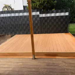 Nowo wybudowany taras z desek kompozytowych w odcieniach brązu, widoczne fragmenty konstrukcji i czarne ogrodzenie panelowe w tle.