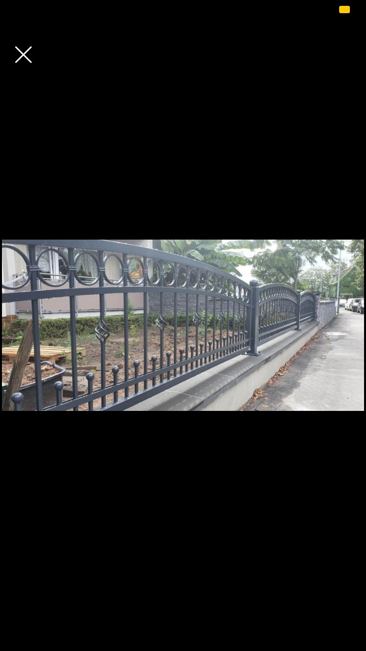 Czarne, metalowe ogrodzenie z dekoracyjnymi elementami w kształcie spirali i okręgów, zamontowane na betonowym murku, widok z boku, w tle roślinność.