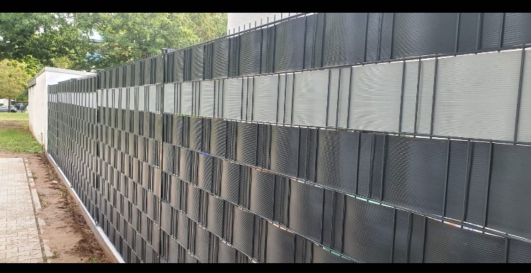 Długi, szary płot z paneli z widoczną strukturą siatki, częściowo wypełniony jasnymi elementami, ciągnący się wzdłuż chodnika i trawnika, z budynkiem w tle.