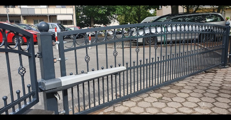 Otwarta, szara, metalowa brama wjazdowa z automatycznym napędem, widok z zewnątrz na podjazd z kostki brukowej i zaparkowane samochody w tle.