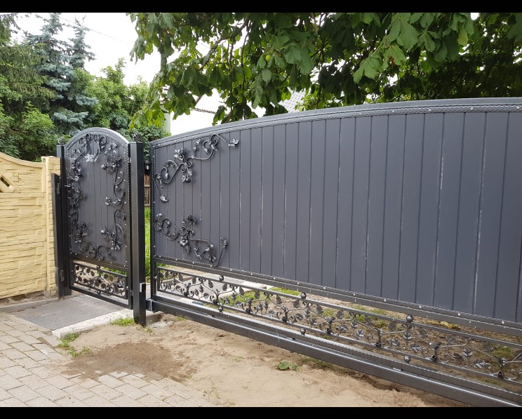 Grafitowa brama wjazdowa z kutymi elementami roślinnymi, widok z zewnątrz posesji, na pierwszym planie kałuża na chodniku.