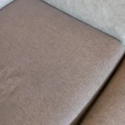 Fragment brązowej tapicerki kanapy z widoczną strukturą materiału po czyszczeniu, z szarym elementem w tle.
