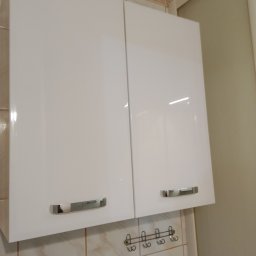 Filip Jagodziński - Białe, lakierowane szafki kuchenne wiszące z chromowanymi uchwytami na tle beżowych płytek i wieszaka na akcesoria kuchenne.