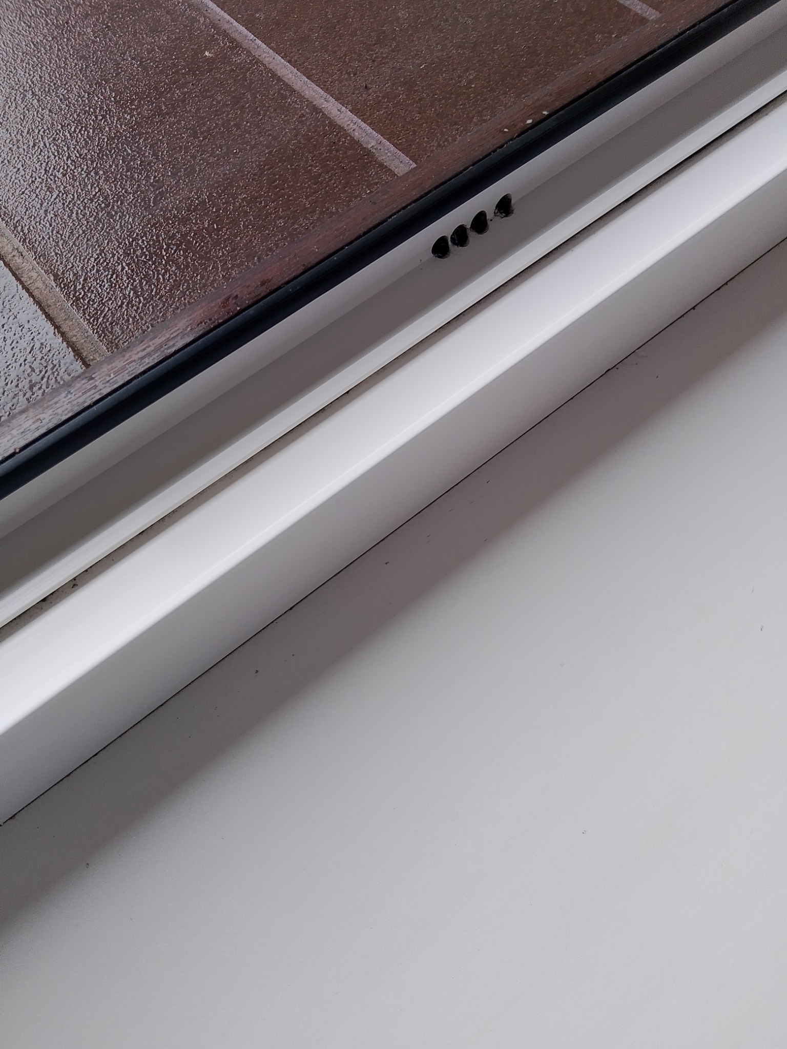 Fragment okna z brązową szybą o fakturze i białym profilem z widocznymi otworami wentylacyjnymi.