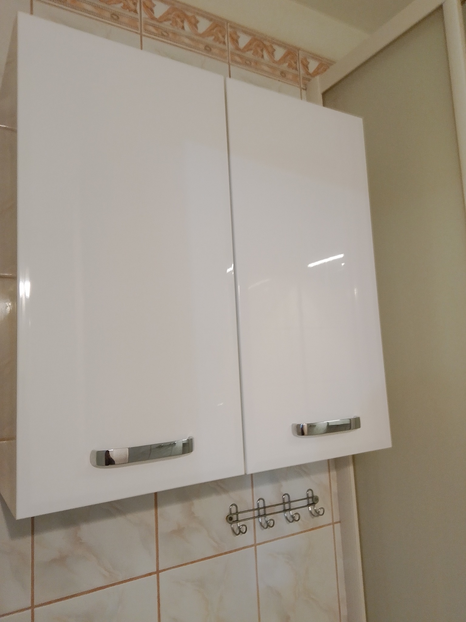 Białe, lakierowane szafki kuchenne wiszące z chromowanymi uchwytami na tle beżowych płytek i wieszaka na akcesoria kuchenne.