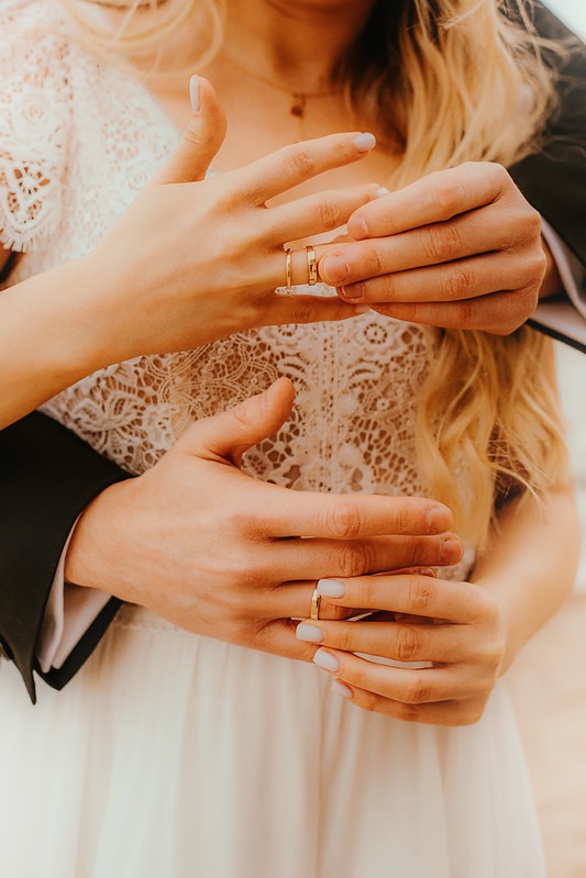 Dłonie nowożeńców podczas ceremonii ślubnej, wymiana złotych obrączek na tle koronkowej sukni ślubnej i czarnego garnituru.