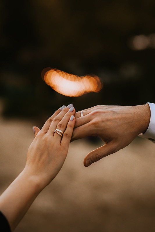 Dłonie pary z obrączkami na tle rozmytego światła, delikatne pastelowe paznokcie kobiety, subtelny gest bliskości.