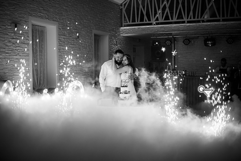 Para młoda obejmuje się za tortem na tle sztucznych ogni i dekoracji świetlnych w sali weselnej z kamienną ścianą.